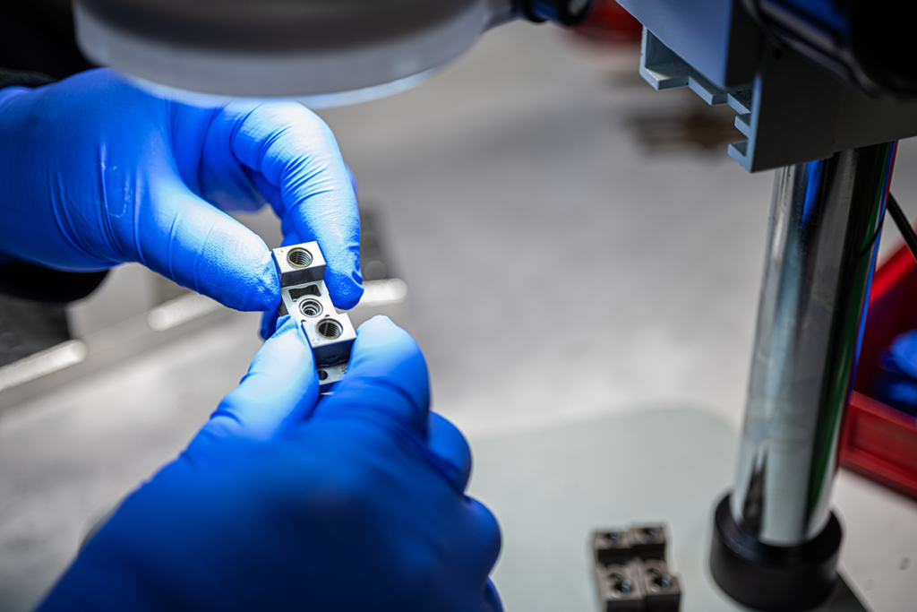 Close-up of a technician in blue nitrile gloves using a microscope to inspect or assemble a small, high-precision metal medical device component.