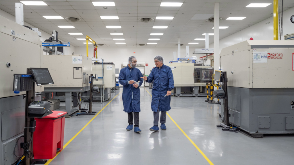 Two male technicians in blue lab coats and hairnets walking and talking through a clean, modern medical device manufacturing facility with large injection molding machines.