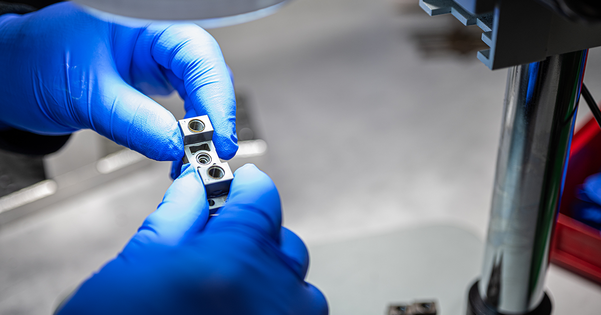 Close-up of a technician in blue nitrile gloves using a microscope to inspect or assemble a small, high-precision metal medical device component.