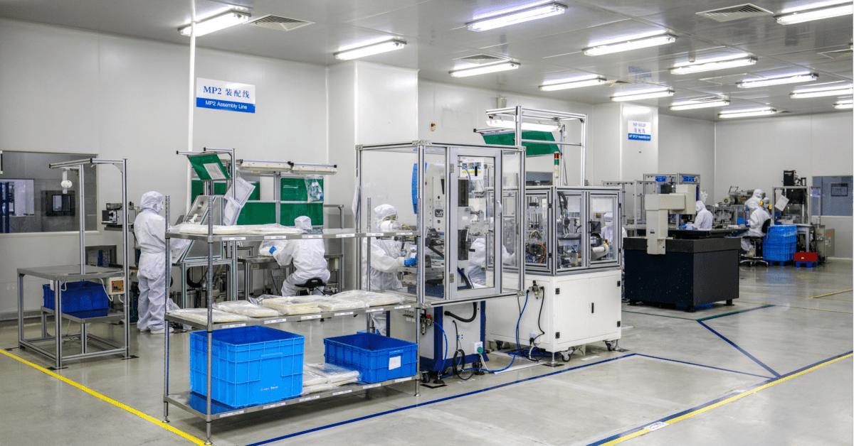 Workers in full white protective suits and headgear operating machinery and organizing components at the MP2 Assembly Line in a clean, brightly lit medical manufacturing facility.